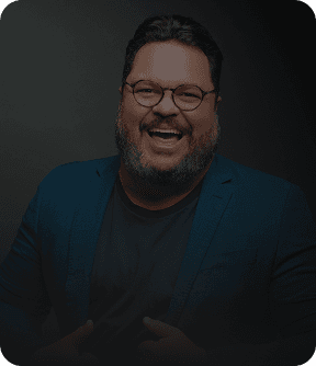 Um painel digital em cinza e preto com um homem sorridente de óculos, barbudo e camisa social azul. Ele aponta para o centro do painel, onde há vários cartões com textos como 'Equipe engajada', 'Clientes agendados' e 'Meta de vendas batida!'. Na parte inferior, há botões com os textos 'Ligar para cliente', 'Chamar no WhatsApp', 'Agendar tarefa', 'Registrar novo contato' e 'Ver convite'.