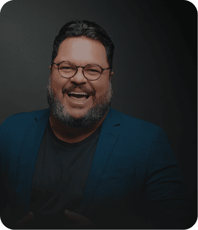 Um painel digital em cinza e preto com um homem sorridente de óculos, barbudo e camisa social azul. Ele aponta para o centro do painel, onde há vários cartões com textos como 'Equipe engajada', 'Clientes agendados' e 'Meta de vendas batida!'. Na parte inferior, há botões com os textos 'Ligar para cliente', 'Chamar no WhatsApp', 'Agendar tarefa', 'Registrar novo contato' e 'Ver convite'.