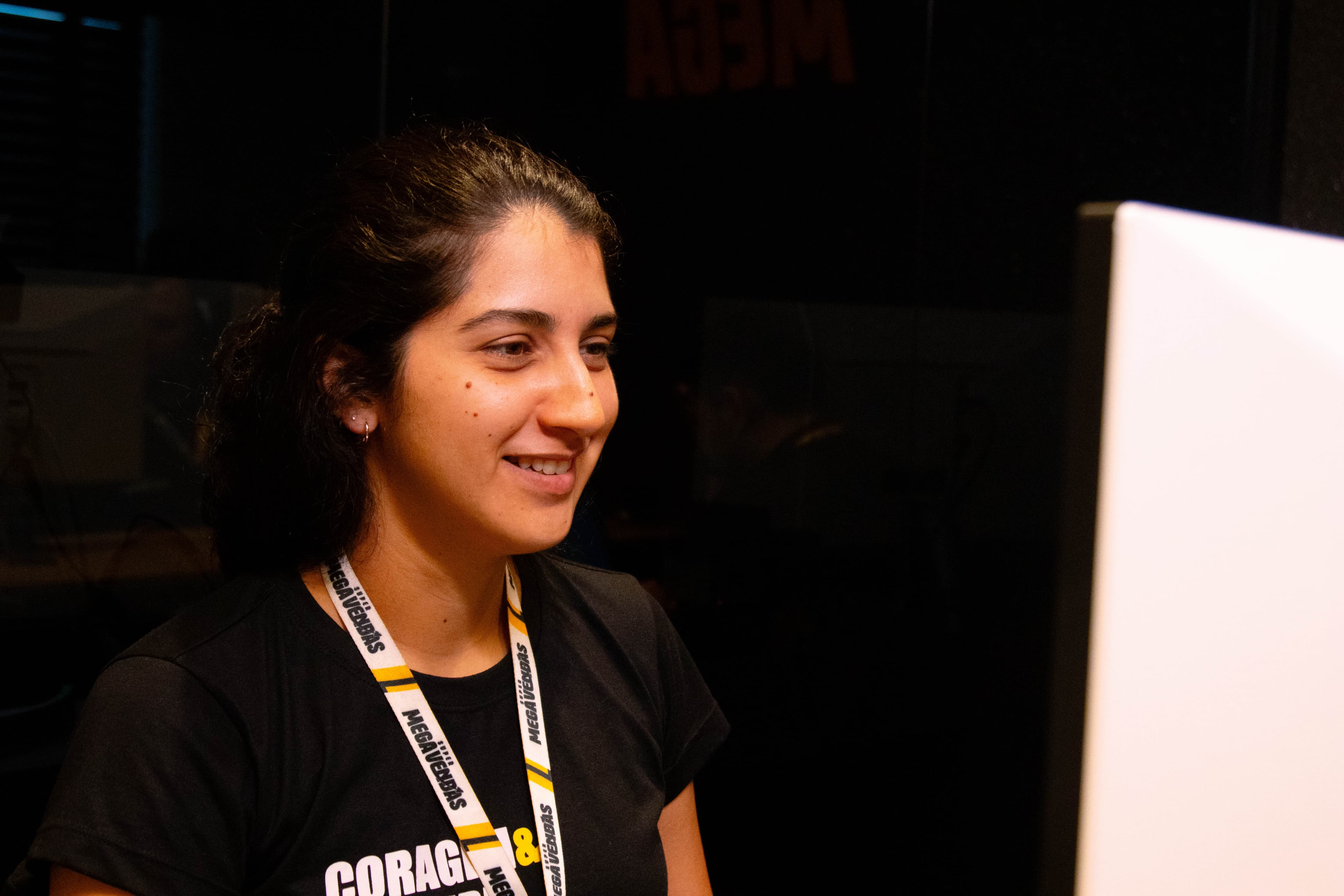 Colaboradora da Super Megavendas sorrindo enquanto trabalha no computador, usando crachá e camiseta da empresa.